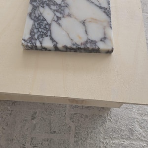 May include: A square marble tile with a white and gray pattern. The tile is set on a light-colored wooden surface. The marble has a polished finish and is likely for use in home decor or construction.