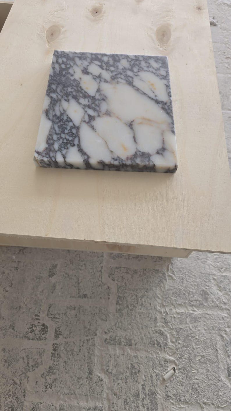 May include: A square marble tile with a white and gray pattern. The tile is set on a light-colored wooden surface. The marble has a polished finish and is likely for use in home decor or construction.