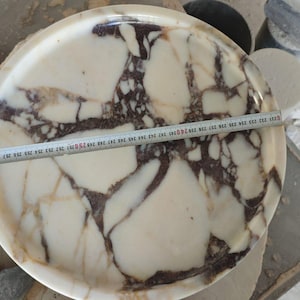 May include: A round, marble tray with a cream and brown patterned design. A measuring tape is laid across the tray, indicating a diameter of approximately 10 inches. The tray is on a work surface with tools and other marble pieces.