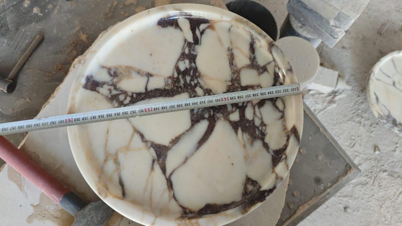 May include: A round, marble tray with a cream and brown patterned design. A measuring tape is laid across the tray, indicating a diameter of approximately 10 inches. The tray is on a work surface with tools and other marble pieces.