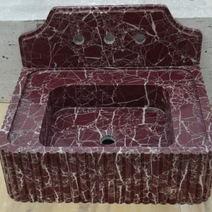 May include: A burgundy marble sink with white veining. The sink has a rectangular basin and a decorative backsplash with three faucet holes. The front of the sink has a ribbed design.