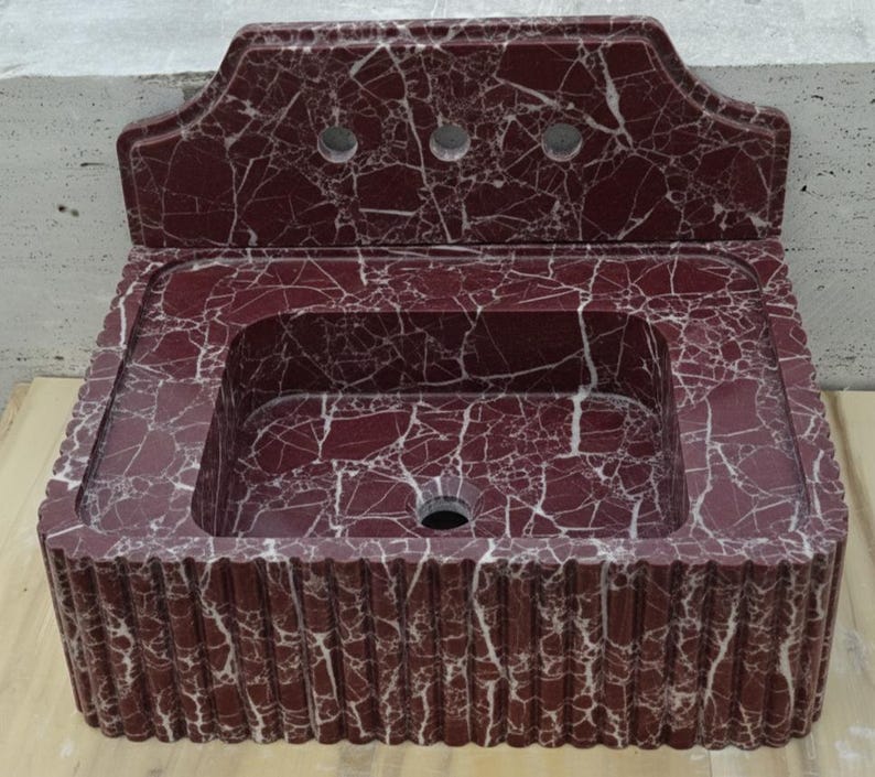 May include: A burgundy marble sink with white veining. The sink has a rectangular basin and a decorative backsplash with three faucet holes. The front of the sink has a ribbed design.