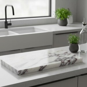 May include: A rectangular marble cutting board with white and burgundy veining sits on a kitchen countertop. A clear glass bottle with a metal spout and a small potted plant are next to the board. A sink and window are in the background.