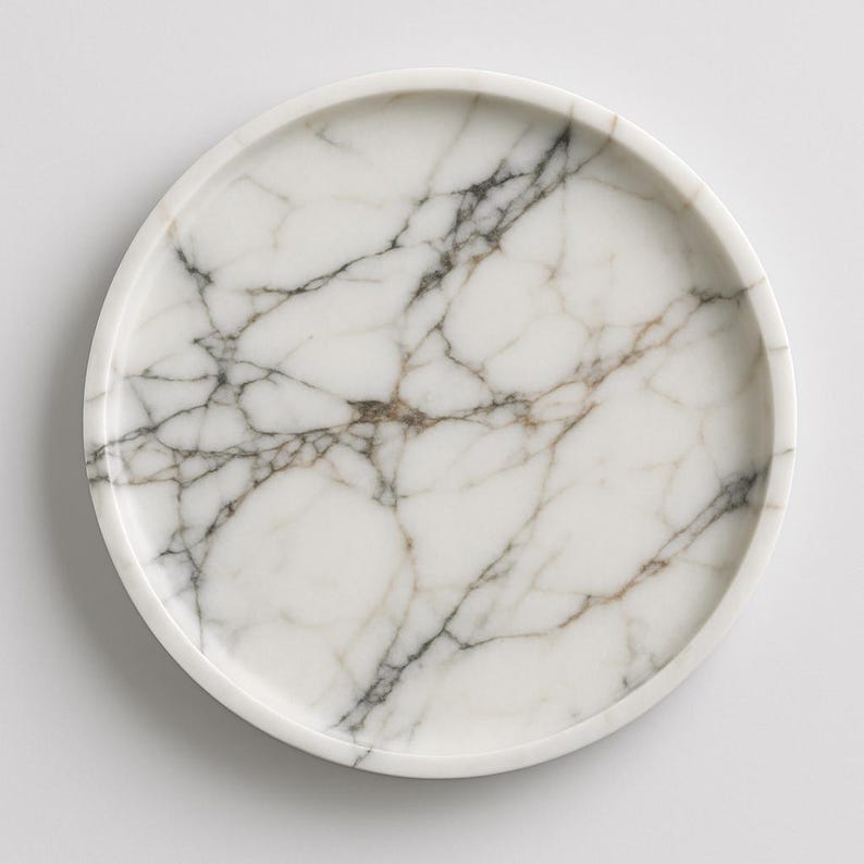 May include: A round, white marble tray with a raised edge. The tray features a natural veining pattern in shades of gray and brown, creating a unique and elegant design. The tray is suitable for serving or display.