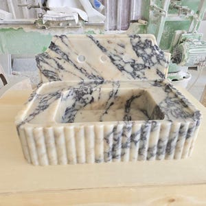 May include: A marble sink with a cream and gray pattern. The sink has a rectangular basin and a backsplash with two holes. The front of the sink has vertical ridges. The sink is sitting on a light-colored wooden surface.