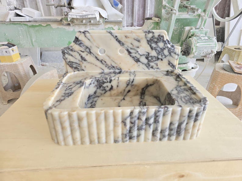 May include: A marble sink with a cream and gray pattern. The sink has a rectangular basin and a backsplash with two holes. The front of the sink has vertical ridges. The sink is sitting on a light-colored wooden surface.