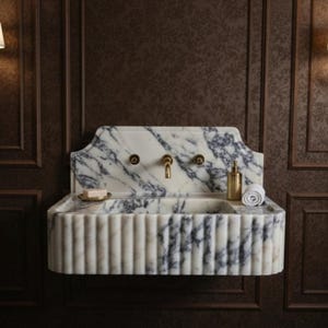May include: A marble bathroom sink with a fluted design and brass fixtures. The sink has a white and gray veined pattern. A small soap dish, a rolled towel, and a gold-colored bottle are on the sink's surface. The sink is mounted on a dark brown wall.