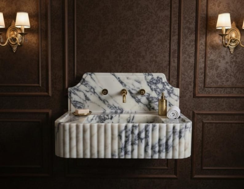 May include: A marble bathroom sink with a fluted design and brass fixtures. The sink has a white and gray veined pattern. A small soap dish, a rolled towel, and a gold-colored bottle are on the sink's surface. The sink is mounted on a dark brown wall.