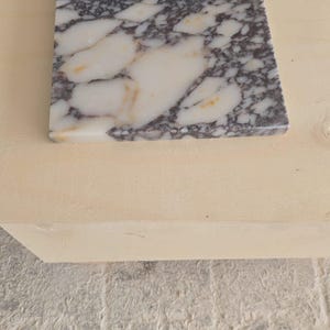 May include: A square marble tile with a white base and dark gray veining. The tile is set on a light-colored wooden surface, with a textured, light gray surface in the background. The marble has a polished finish.