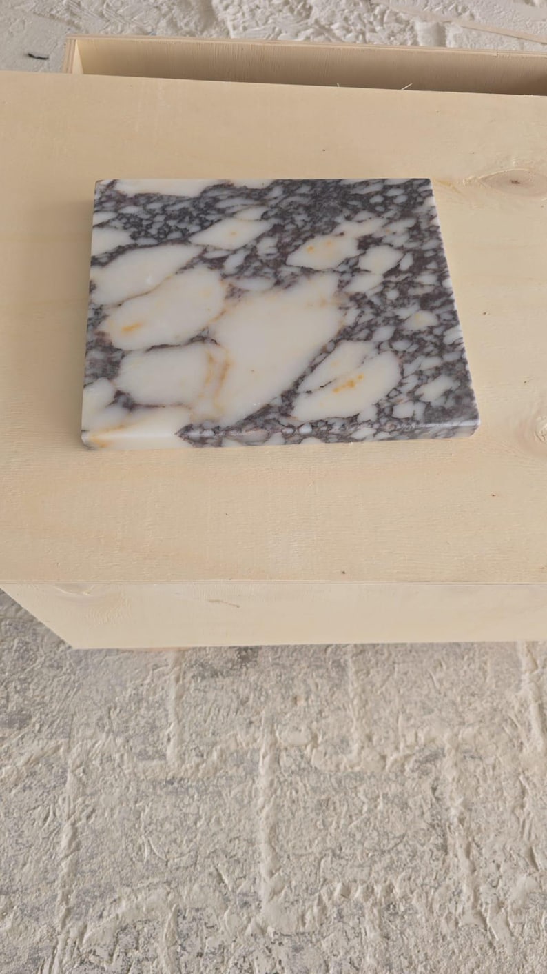 May include: A square marble tile with a white base and dark gray veining. The tile is set on a light-colored wooden surface, with a textured, light gray surface in the background. The marble has a polished finish.