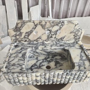 May include: A marble sink with a rectangular basin and a decorative scalloped edge. The sink has a white and gray veined pattern, with three faucet holes. The overall design is ornate and elegant, suitable for a bathroom.