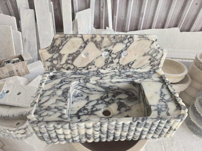 May include: A marble sink with a rectangular basin and a decorative scalloped edge. The sink has a white and gray veined pattern, with three faucet holes. The overall design is ornate and elegant, suitable for a bathroom.