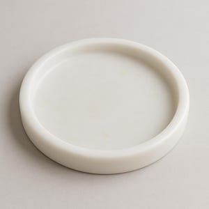 May include: A round, white marble tray with a slightly raised edge. The tray has a smooth, polished surface and a minimalist design. Ideal for serving or displaying small items. The tray is approximately 6 inches in diameter.