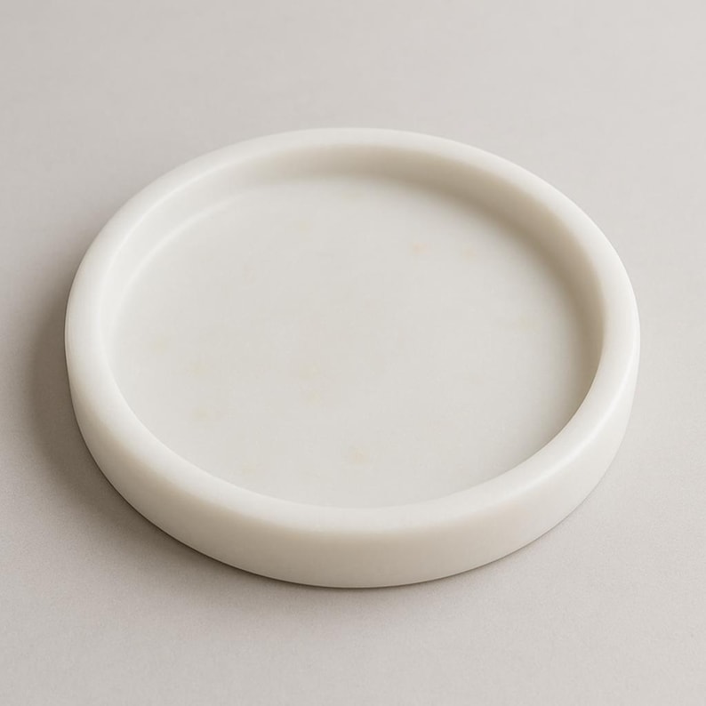 May include: A round, white marble tray with a slightly raised edge. The tray has a smooth, polished surface and a minimalist design. Ideal for serving or displaying small items. The tray is approximately 6 inches in diameter.