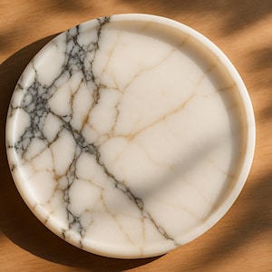 Pode incluir: Uma bandeja redonda de mármore branco sujo com uma borda. Apresenta um padrão marcante de veios cinzentos e dourados. A bandeja está colocada sobre uma superfície de madeira, com sombras projetadas sobre a madeira.