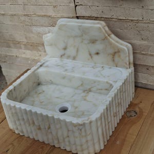 May include: A white marble sink with a decorative back and a fluted exterior. The sink has a rectangular basin with a central drain hole. The marble has brown veining throughout, and it sits on a wooden surface.