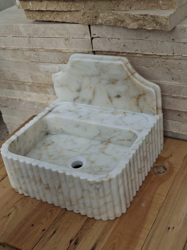 May include: A white marble sink with a decorative back and a fluted exterior. The sink has a rectangular basin with a central drain hole. The marble has brown veining throughout, and it sits on a wooden surface.