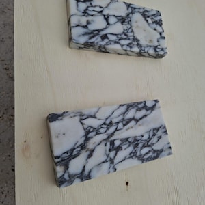 May include: Two rectangular marble tiles with a white base and intricate black veining. The tiles are positioned on a light-colored wooden surface, showcasing their natural stone patterns and textures. The marble's polished surface reflects light, highlighting the contrast.