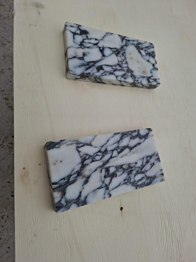 May include: Two rectangular marble tiles with a white base and intricate black veining. The tiles are positioned on a light-colored wooden surface, showcasing their natural stone patterns and textures. The marble's polished surface reflects light, highlighting the contrast.