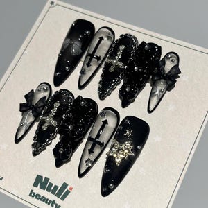Puede incluir: Un conjunto de uñas postizas negras y plateadas con varios diseños, incluyendo cruces, lazos y estrellas. Algunas uñas están adornadas con pedrería y purpurina. Las uñas se muestran en una tarjeta blanca con el texto "Nuli beauty".