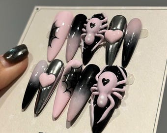 May include: A set of ten long, pointed false nails. The nails feature a variety of designs, including pink and black ombre, spiderweb graphics, and 3D pink spiders and hearts. The brand name "Nuli beauty" is visible.