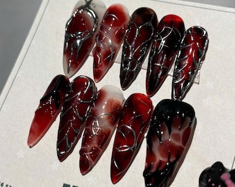 May include: A set of ten long, pointed artificial nails. Each nail features a gradient design, transitioning from a clear or white base to a deep red or black tip. Silver metallic accents add a decorative touch. The nails are arranged on a white surface.