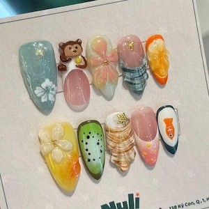 May include: A set of ten artificial nails with diverse designs. The nails showcase a monkey, flowers, fruit, and a fish. The colour palette includes blue, pink, yellow, green, and orange. Some nails have 3D decorations.