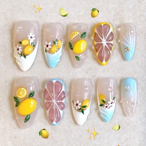 May include: A collection of ten artificial nails with fruit-themed designs. The nails feature yellow lemons, grapefruit slices, and floral accents. The nails have a clear base with white, blue, and yellow details, some with a 3D effect.