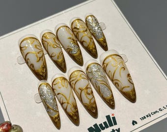May include: A set of ten long, almond-shaped press-on nails. Each nail features a white base with gold floral designs and gold tips. Some nails have silver glitter accents. The brand name "Nuli Beauty" is visible.