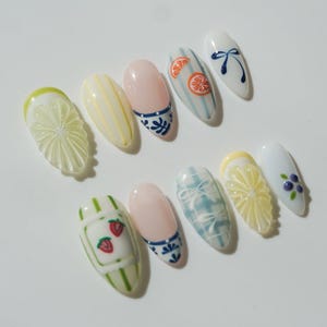 May include: A collection of painted artificial nails with various designs. The nails feature fruit slices, stripes, floral patterns, and bows in shades of white, yellow, pink, and blue. The nails are oval-shaped.