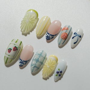 May include: A collection of painted artificial nails with various designs. The nails feature fruit slices, strawberries, floral patterns, and bow accents. Colors include white, pink, blue, yellow, and green. The nails are oval-shaped and have a glossy finish.