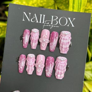 May include: A set of ten pink press-on nails. Some nails have a seashell design, others feature a flower accent, and some are decorated with glitter. The black card behind the nails reads "NAIL BOX for you."