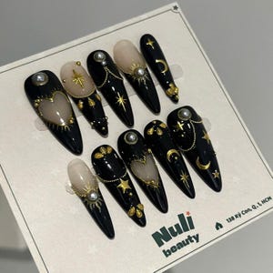 Peut inclure: Un ensemble de dix faux ongles longs, noirs, en forme d'amande. Chaque ongle présente des motifs célestes dorés, notamment des étoiles, des lunes et des soleils, avec des accents de perles. Certains ongles ont une base couleur chair avec des ornements dorés.