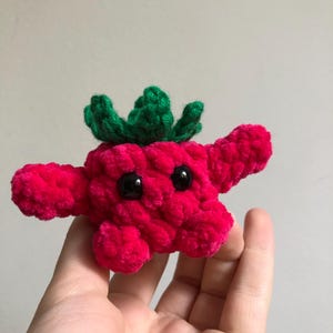 Pode incluir: Uma criatura vermelha de pelúcia feita à mão com folhas verdes no topo e olhos pretos. O brinquedo macio e fofo tem braços pequenos e um design caprichoso, ideal para uma coleção de brinquedos infantis.