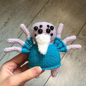 May include: A crocheted spider toy with a light purple body, eight legs, and multiple black eyes. It is dressed in a turquoise blue shirt and has a white beard. The toy is held in a person's hand.