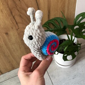 May include: A crocheted snail toy in grey yarn. The shell has a blue and pink spiral pattern. The snail has black eyes and two small antennae. The toy is held in a hand, with a plant in the background.