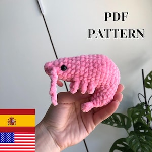 May include: A pink crochet shrimp with black eyes and detailed legs. The image includes the text "PDF PATTERN". The shrimp is held in a hand, with a Spanish and American flag in the corner.