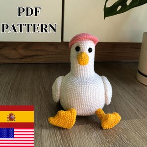 Puede incluir: Un pato blanco tejido a ganchillo con un sombrero rosa, pico y patas amarillas. El pato tiene ojos negros y está sobre una superficie de madera. El texto "PDF PATTERN" es visible. Es un artículo artesanal.