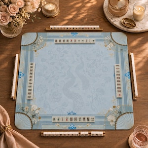 May include: A square mahjong mat in light blue featuring a subtle floral and peacock pattern with fan-shaped corner designs. Four wooden racks hold white mahjong tiles with black, red, and green characters, symbols, and numbers arranged on the mat.
