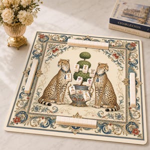May include: A square mahjong mat featuring a cream background with a central illustration of two cheetahs flanking a potted topiary. The border displays ornate blue and red floral scrollwork. Several white mahjong tiles are placed on the mat.