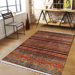 Può includere: Un tappeto rettangolare con un motivo geometrico nei toni del rosso, arancione, blu e marrone. Il tappeto ha un design a righe con una frangia bianca lungo i bordi. È posizionato su un pavimento in legno.