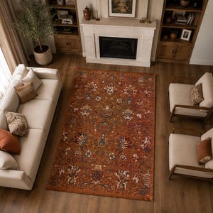 May include: A rectangular area rug featuring a deep burnt orange background with an intricate, symmetrical floral and botanical pattern in shades of blue, cream, and dark brown, centered in a living room with light-colored furniture and wood floors.