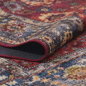 K&ouml;nnte beinhalten: Nahaufnahme eines aufgerollten Teppichs im persischen Stil. Der Teppich zeigt ein traditionelles Design mit einer Farbpalette aus Rot, Blau und Gold. Er hat komplizierte Muster und eine dunkelgraue R&uuml;ckseite. Die Kanten sind mit einem roten Rand versehen.