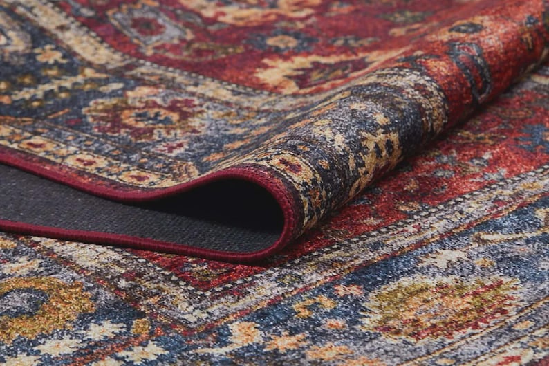 K&ouml;nnte beinhalten: Nahaufnahme eines aufgerollten Teppichs im persischen Stil. Der Teppich zeigt ein traditionelles Design mit einer Farbpalette aus Rot, Blau und Gold. Er hat komplizierte Muster und eine dunkelgraue R&uuml;ckseite. Die Kanten sind mit einem roten Rand versehen.