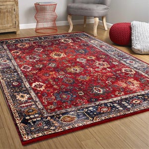 K&ouml;nnte beinhalten: Ein rechteckiger Teppich mit traditionellem Design. Der Teppich hat ein tiefrotes Mittelfeld mit Blumenmustern in Creme, Blau und Gold. Ein dunkelblauer Rand umrahmt das Design. Der Teppich liegt auf einem Holzboden.