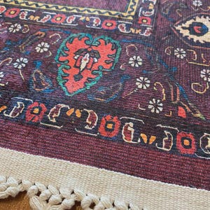 May include: Close-up of a burgundy rug with intricate patterns in red, yellow, and blue. The rug features a central floral design and a cream-colored fringe with tassels. The design is reminiscent of traditional oriental rugs.