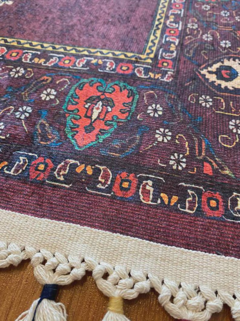 May include: Close-up of a burgundy rug with intricate patterns in red, yellow, and blue. The rug features a central floral design and a cream-colored fringe with tassels. The design is reminiscent of traditional oriental rugs.