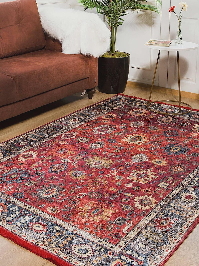 K&ouml;nnte beinhalten: Ein rechteckiger, roter Teppich mit einem komplizierten, traditionellen Muster. Der Teppich hat ein rotes Mittelfeld mit einem detaillierten Blumendesign in Gold-, Blau- und Cremet&ouml;nen. Ein dunkelblauer Rand rahmt den Teppich ein und sorgt f&uuml;r Kontrast und Definition.