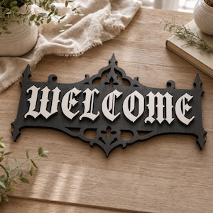 May include: A black wooden sign with the word "WELCOME" in white, gothic-style lettering. The sign has an ornate, decorative border, suitable for home decoration.