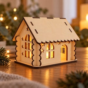 Peut inclure: Une décoration de maison en bois clair avec un bord de toit festonné et des bords brun foncé. La structure présente des fenêtres arquées et des découpes en forme d'étoile, avec une lumière jaune chaude brillant de l'intérieur.